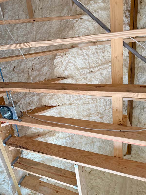 Close-up of a wall under construction with wooden studs and white open-cell spray foam insulation filling the cavities.