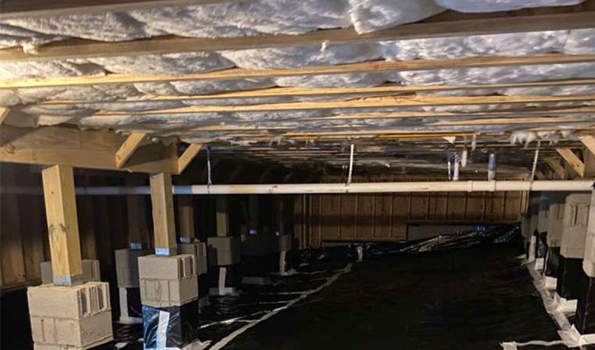 An eye-level shot of a residential crawlspace featuring white fiberglass insulation between ceiling joists, concrete support piers, and a black plastic vapor barrier covering the entire floor.