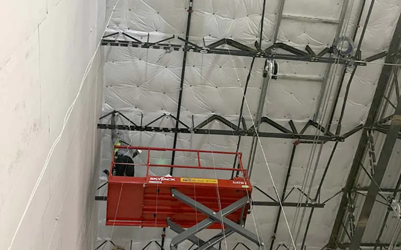A Hayes Insulation installer on a red Skyjack scissor lift securing white vinyl-faced fiberglass insulation to the ceiling of a large metal building in Nashville.