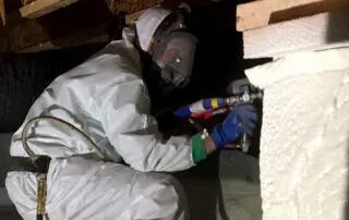 Technician in full protective suit and respirator applying spray foam insulation in a crawl space.