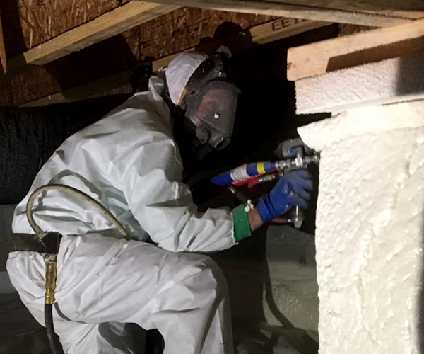 Technician in full protective suit and respirator applying spray foam insulation in a crawl space.
