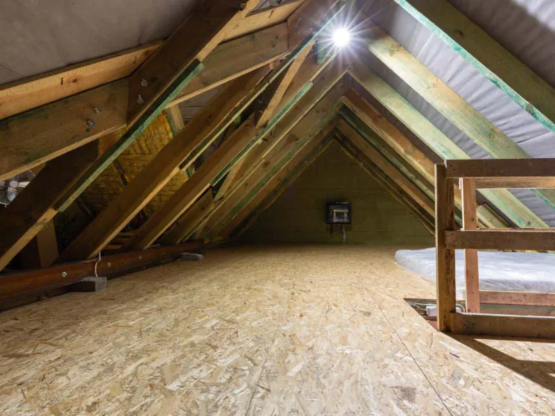 Empty attic interior with exposed wood trusses, OSB subfloor, and a single overhead work light.