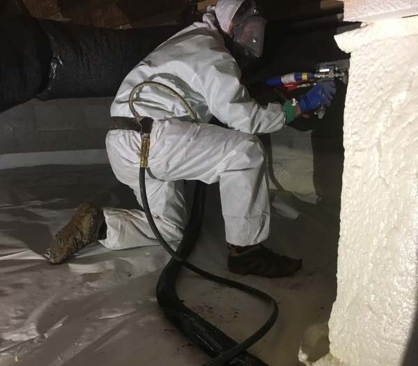 A professional technician from Hayes Insulation in Nashville, wearing full protective gear and a respirator, applies spray foam insulation to seal a crawl space foundation wall over a white vapor barrier.