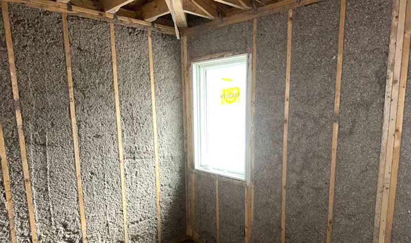 Dense-pack cellulose insulation filling framed wall cavities in a new construction room with a small window.