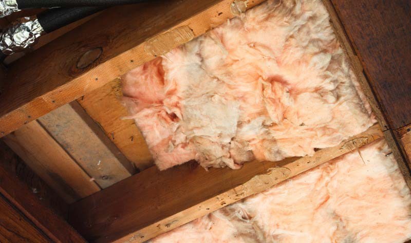 Exposed insulation between wooden joists in ceiling. Discolored fiberglass batt with gaps and uneven coverage causing potential heat loss and poor thermal efficiency. Selective focus.