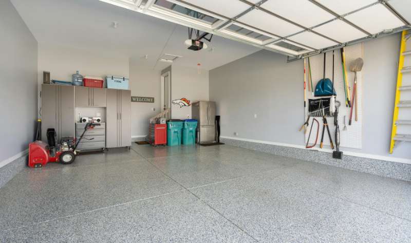 A clean, finished two-car garage with gray epoxy-coated speckled flooring, gray wall-mounted storage cabinets, a red tool chest, a snow blower, pegboard tool organizer with garden tools, a yellow ladder, and a garage door opener rail overhead.