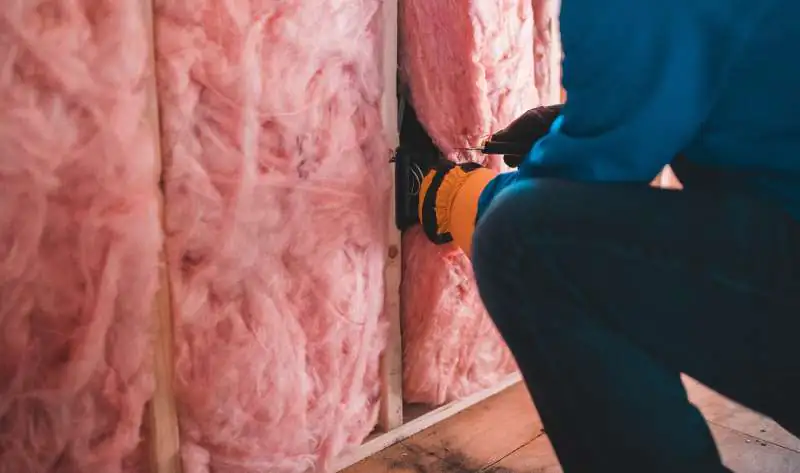 A worker wearing orange gloves and a blue hoodie crouching to install pink fiberglass batt insulation between wooden wall studs during a home construction or renovation project.